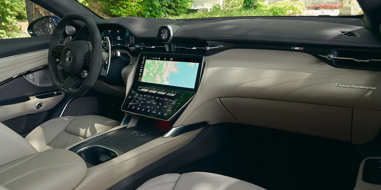 Cockpit des Maserati GranTurismo Modelljahr 2027 mit Lenkrad, Digitaluhr, Zentraldisplay mit Navigationskarte, PRND-Wählschalter und Komfort-Display, Interieur in Hellbeige